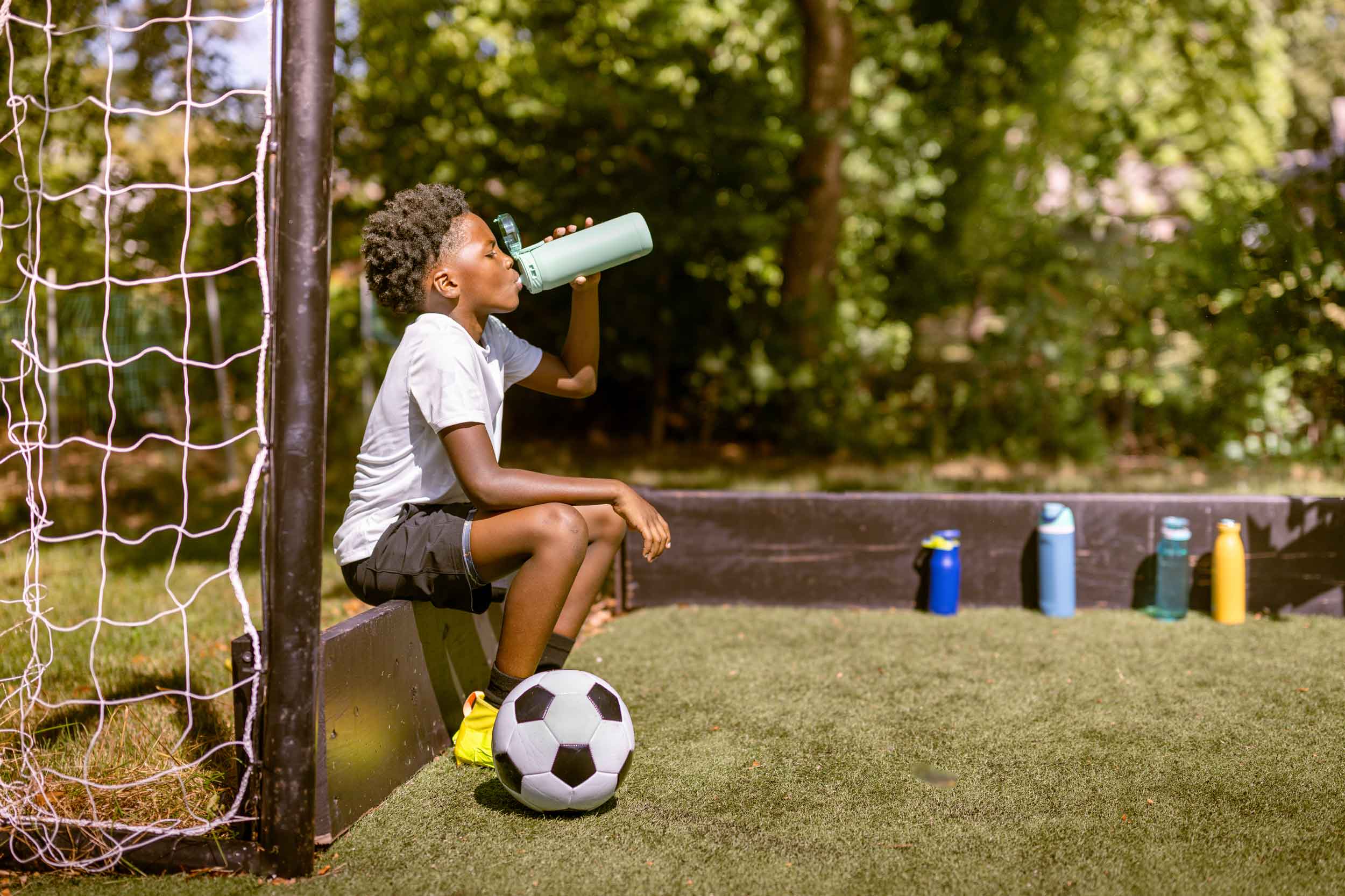 Young child sitting down drinking water form a water bottle with a soccer ball.